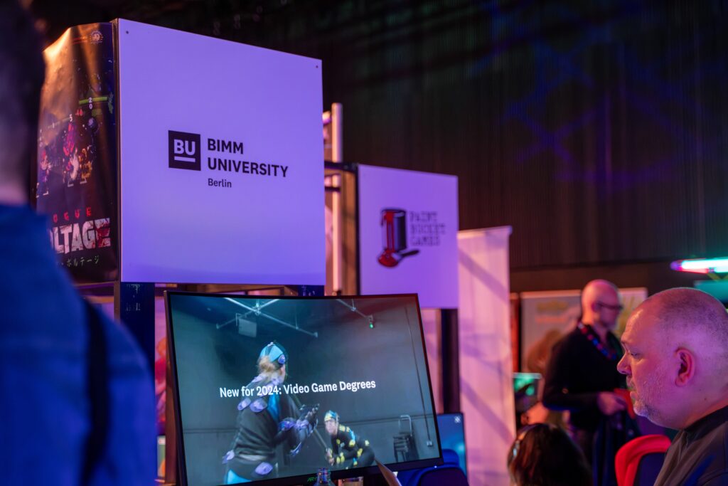 BIMM University's Dean of Creative Technologies Nick Rodriguez stands at BIMM University Berlin's exhibition stand at Games Ground. On a computer screen below the BIMM University Berlin sign reads "New for 2024: Video Game Degrees"